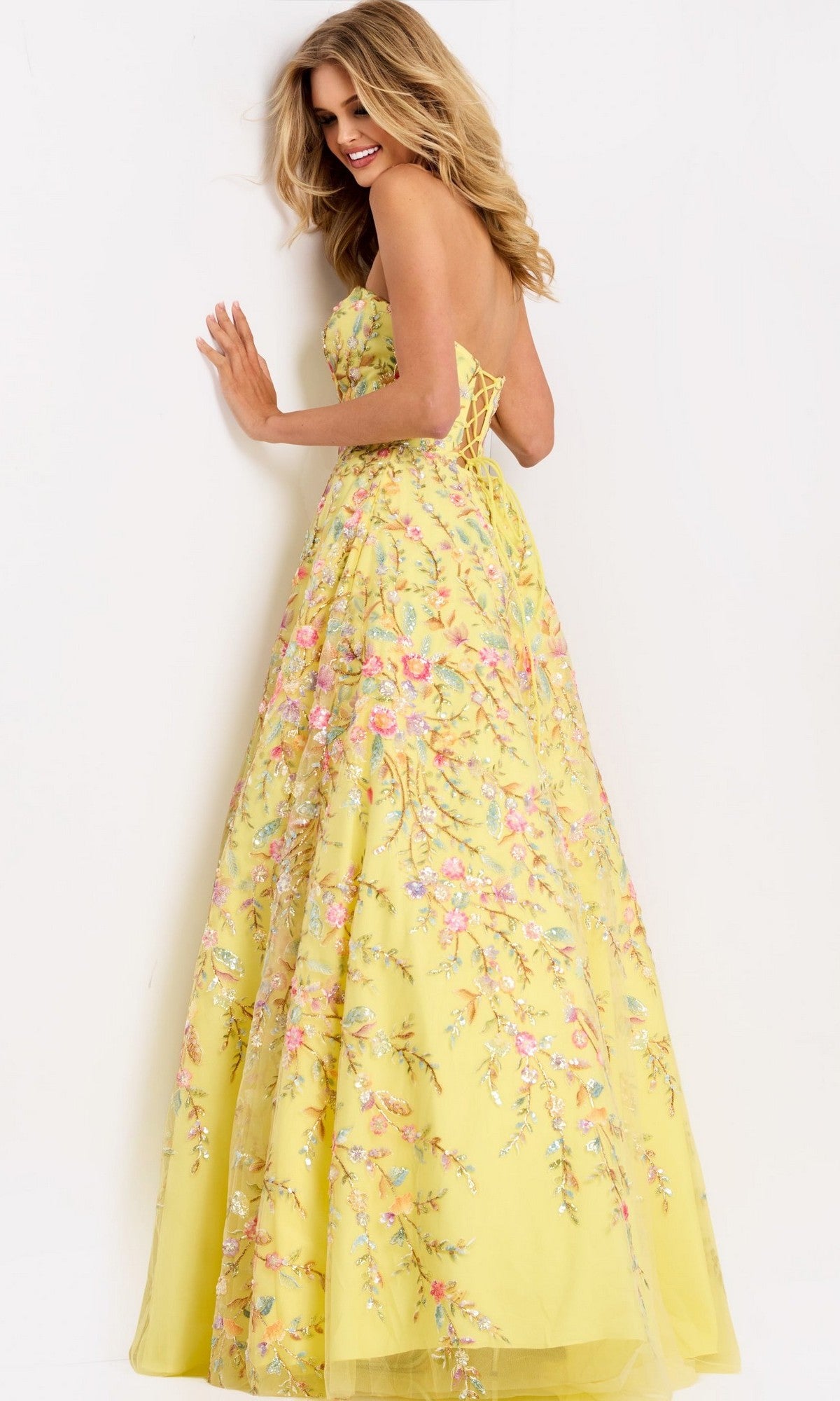 A woman smiles against a white wall while wearing the Jovani 49070 long prom dressโa yellow strapless ball gown featuring floral embroidery.